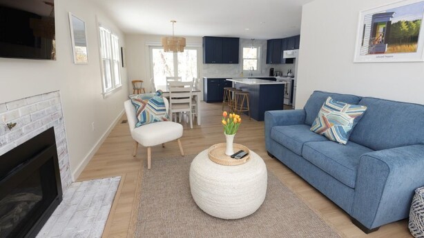 Een moderne woonkamer met een open haard, een blauwe bank, een ronde salontafel met een bloemenvaas, en een keuken op de achtergrond.