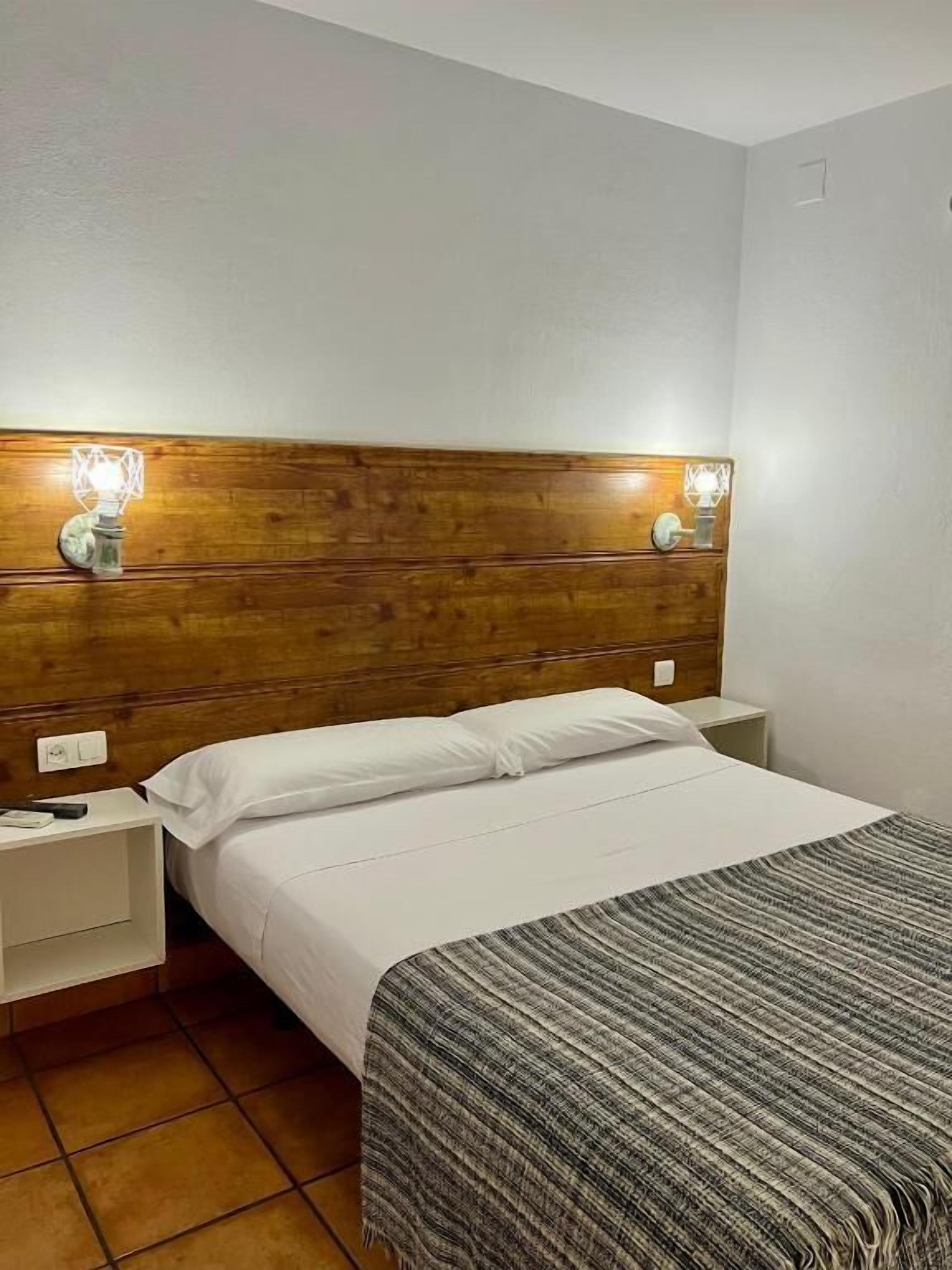Une chambre d’hôtel avec une tête de lit en bois, du linge de lit blanc, un plaid rayé gris et des lampes de chevet.