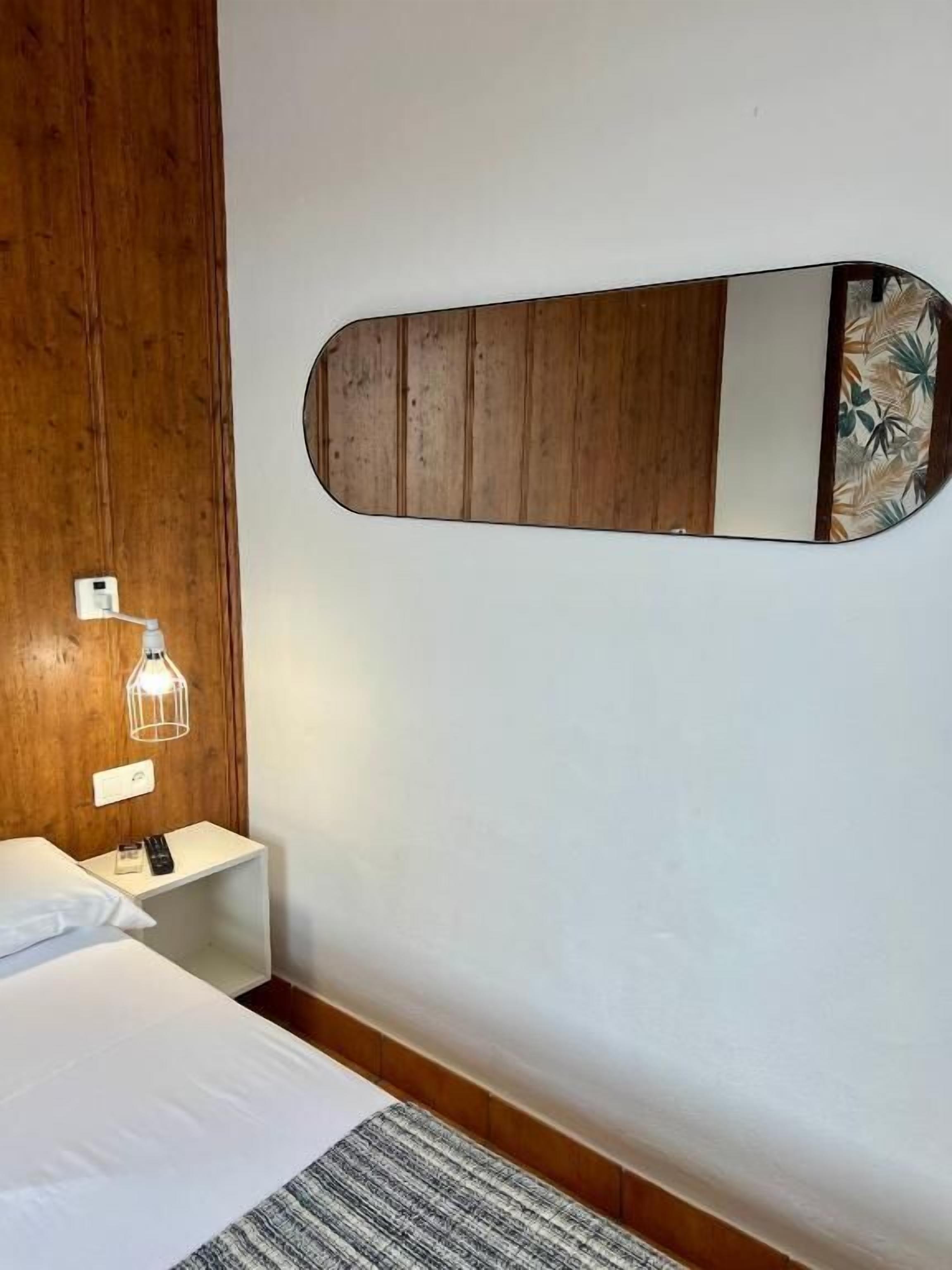 Habitación de hotel con cama, mesita de noche con lámpara, mesita auxiliar y espejo montado en la pared.