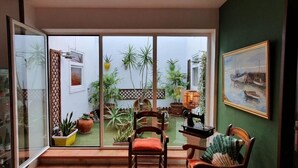 A room with a sliding glass door leading to a garden with potted plants. There is a wooden chair with a cushion, a table with a lamp, and a framed painting on the wall. - Hostal Real (San Juan del Puerto)