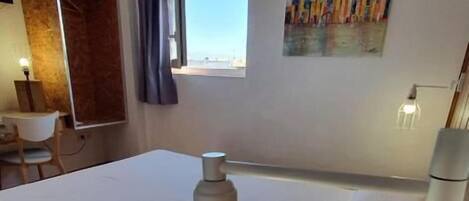 Habitación de hotel con cama, escritorio, silla, ventana con cortinas, un cuadro en la pared y una lámpara colgante.