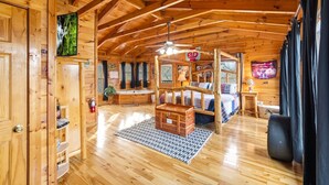 Interior de cabana rústica com teto de madeira, cama com coberta xadrez, baú de madeira e mesa pequena com abajur.