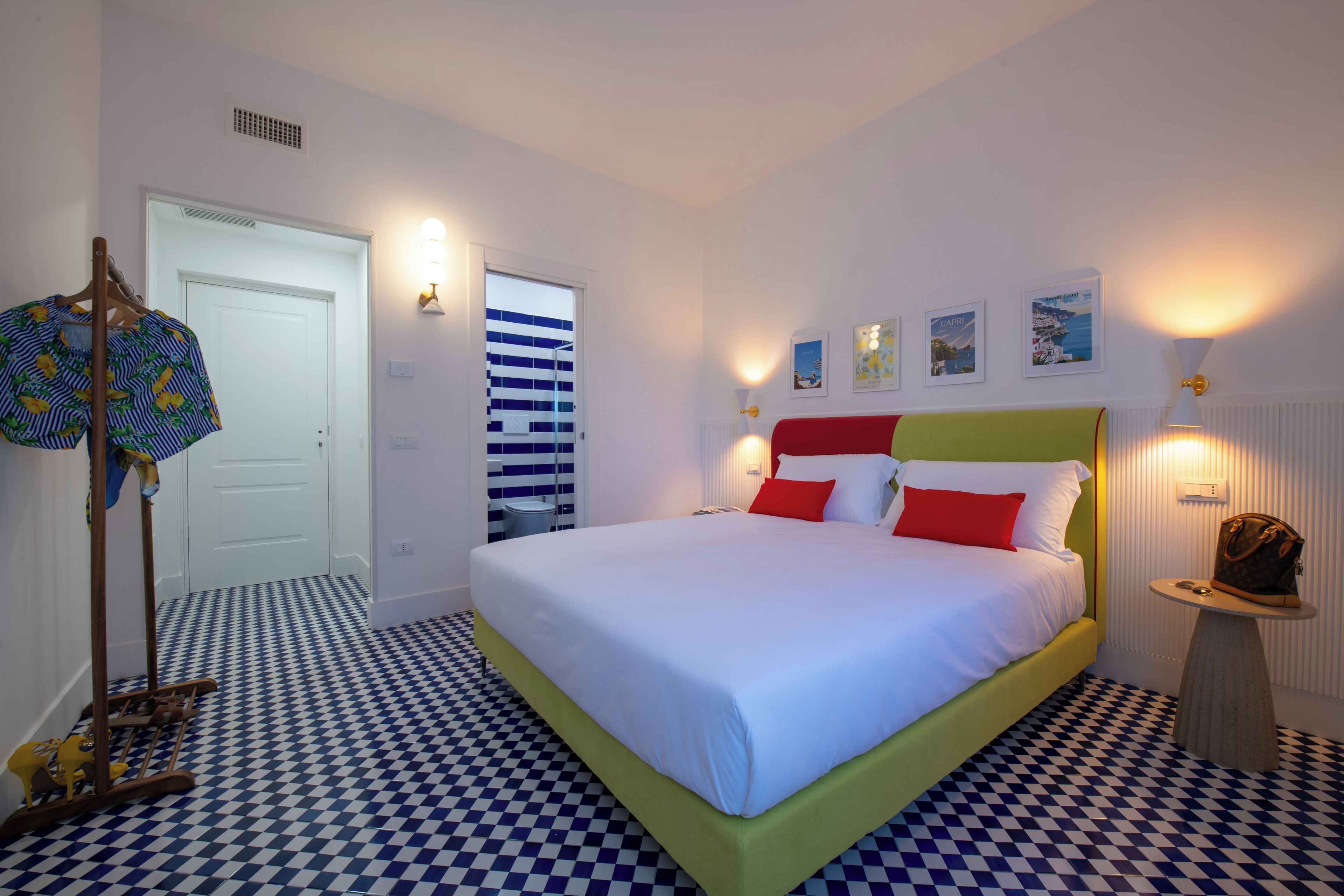 A hotel room with a checkered floor, a bed with white and red bedding, a wooden chair, a small table with a lamp, and a door with a striped curtain.