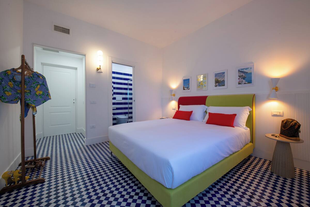 A hotel room with a checkered floor, a bed with white and red bedding, a wooden chair, a small table with a lamp, and a door with a striped curtain.