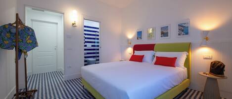Habitación de hotel con piso a cuadros, cama con ropa de cama blanca y roja, una silla de madera, una mesita con lámpara y una puerta con cortina a rayas.