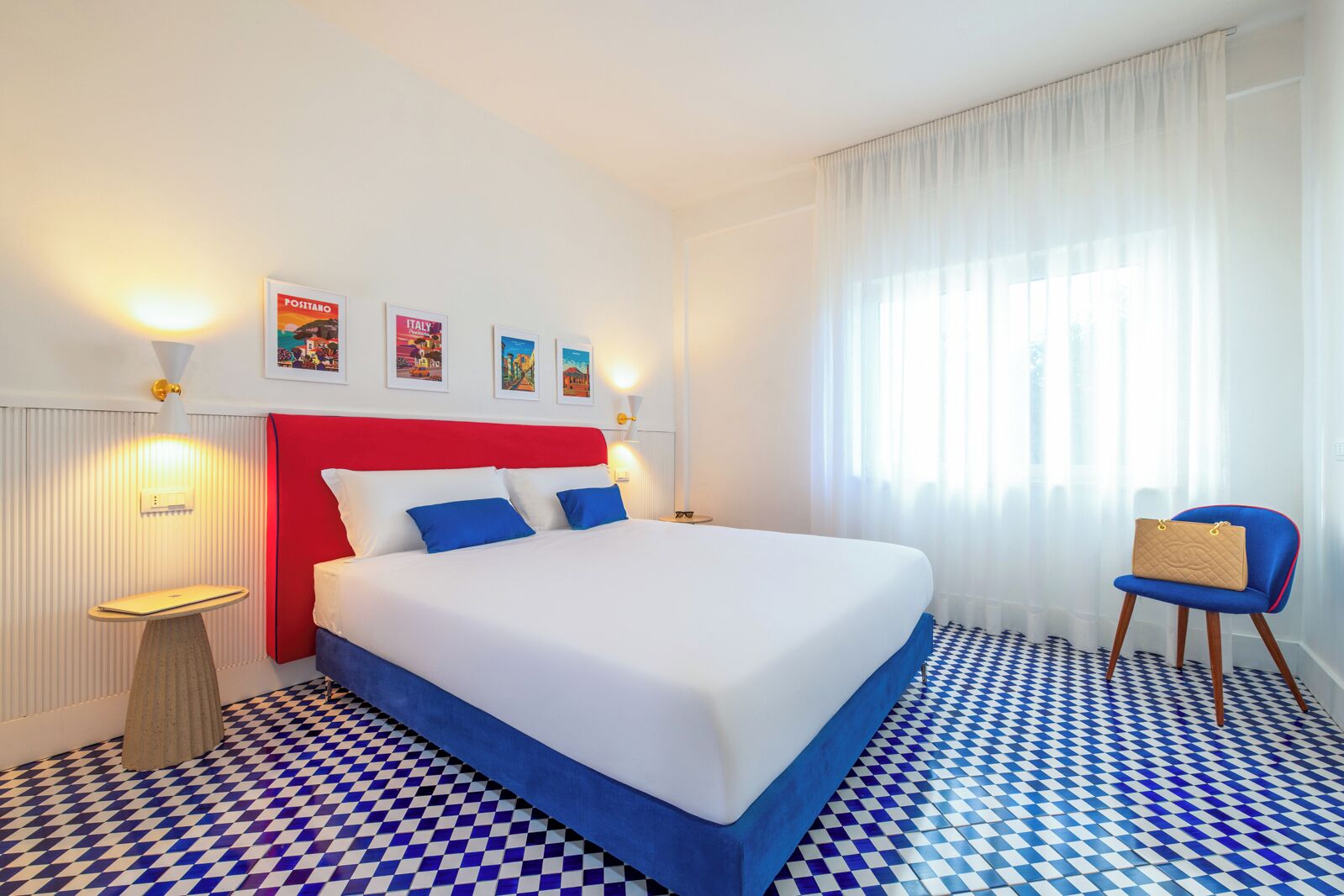 Ein Hotelzimmer mit einem großen Bett, einem roten Kopfteil und einem karierten Boden. An der Wand sind Leuchten befestigt und ein kleiner Tisch mit einer Lampe steht bereit.