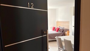 Una habitación moderna con una puerta negra numerada con el número 12, una zona de comedor con sillas blancas y una cama con cabecera y almohadas.
