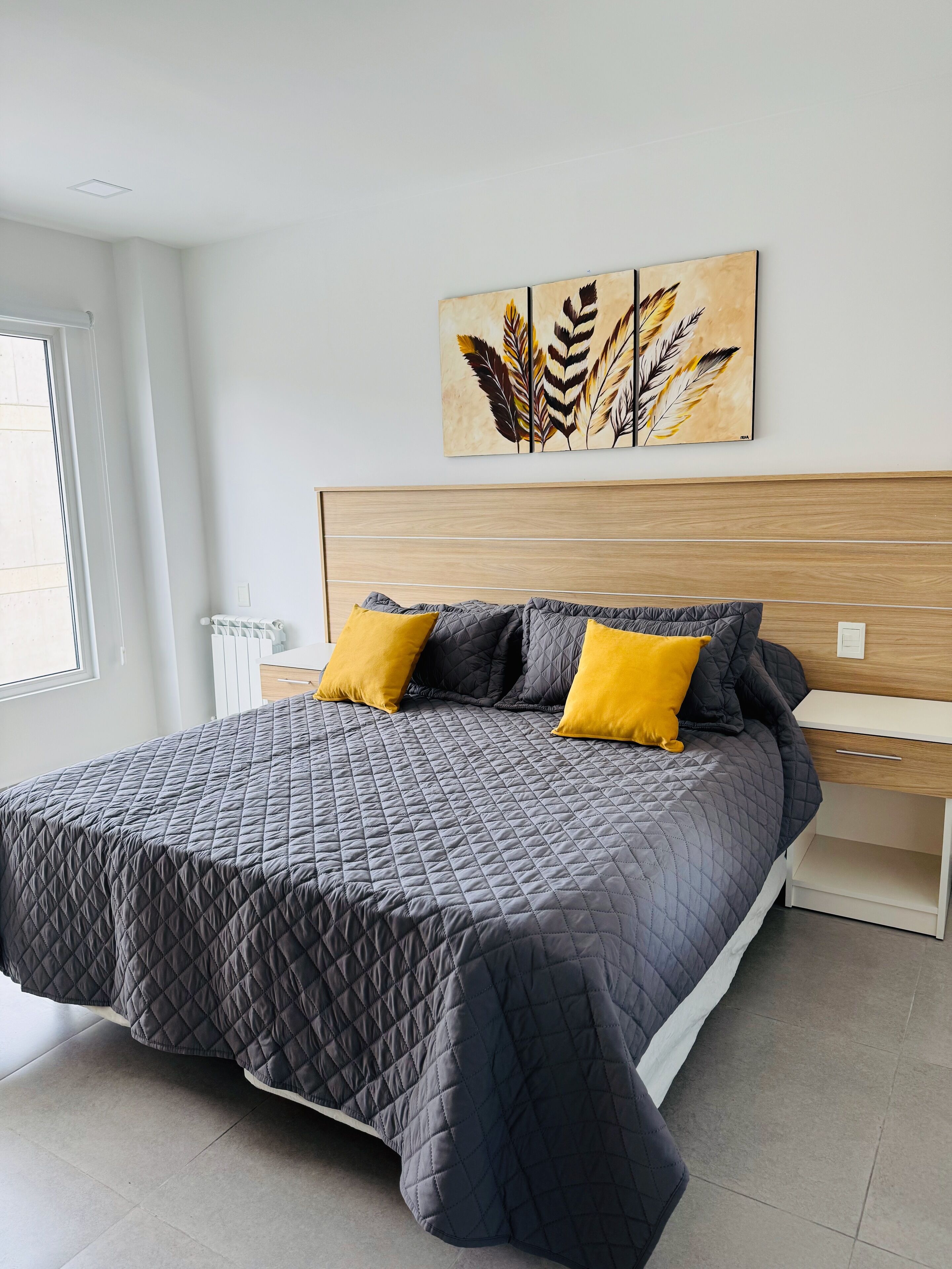 Ein ordentlich bezogenes Bett mit einer grauen Decke, zwei gelben Kissen und einem Holz-Kopfstück.