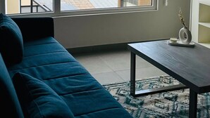 Una sala de estar moderna con un amplio ventanal que ofrece vistas a una masa de agua, un sof á azul, una mesita con un florero y una estantería.
