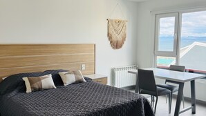 Un dormitorio moderno con una cama grande, cabecera de madera, una mesa de comedor con sillas y vista al mar a través de un ventanal amplio.