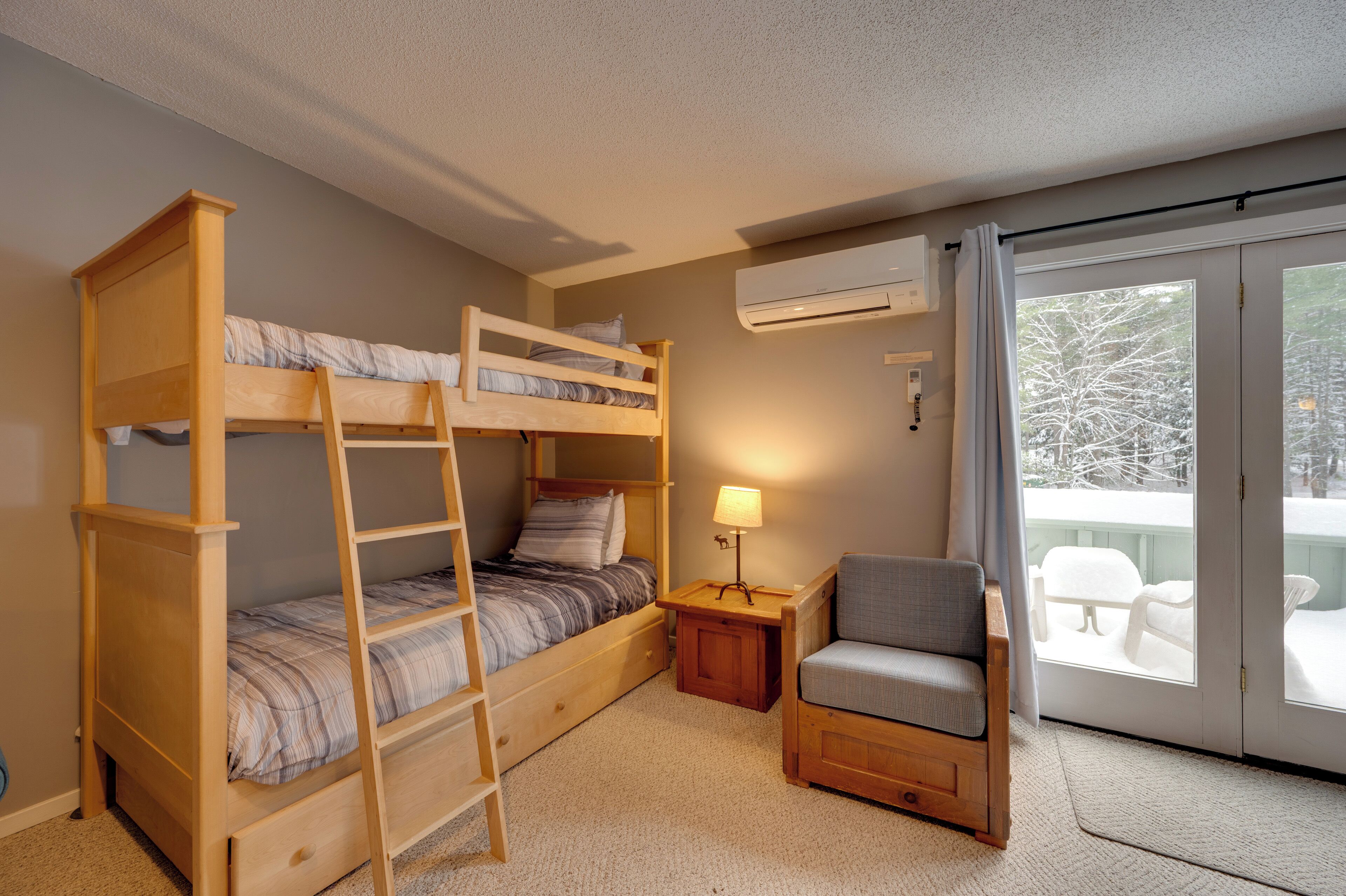 A bunk bed room with a ladder, a small table with a lamp, a chair, and a sliding glass door leading to an outdoor area.