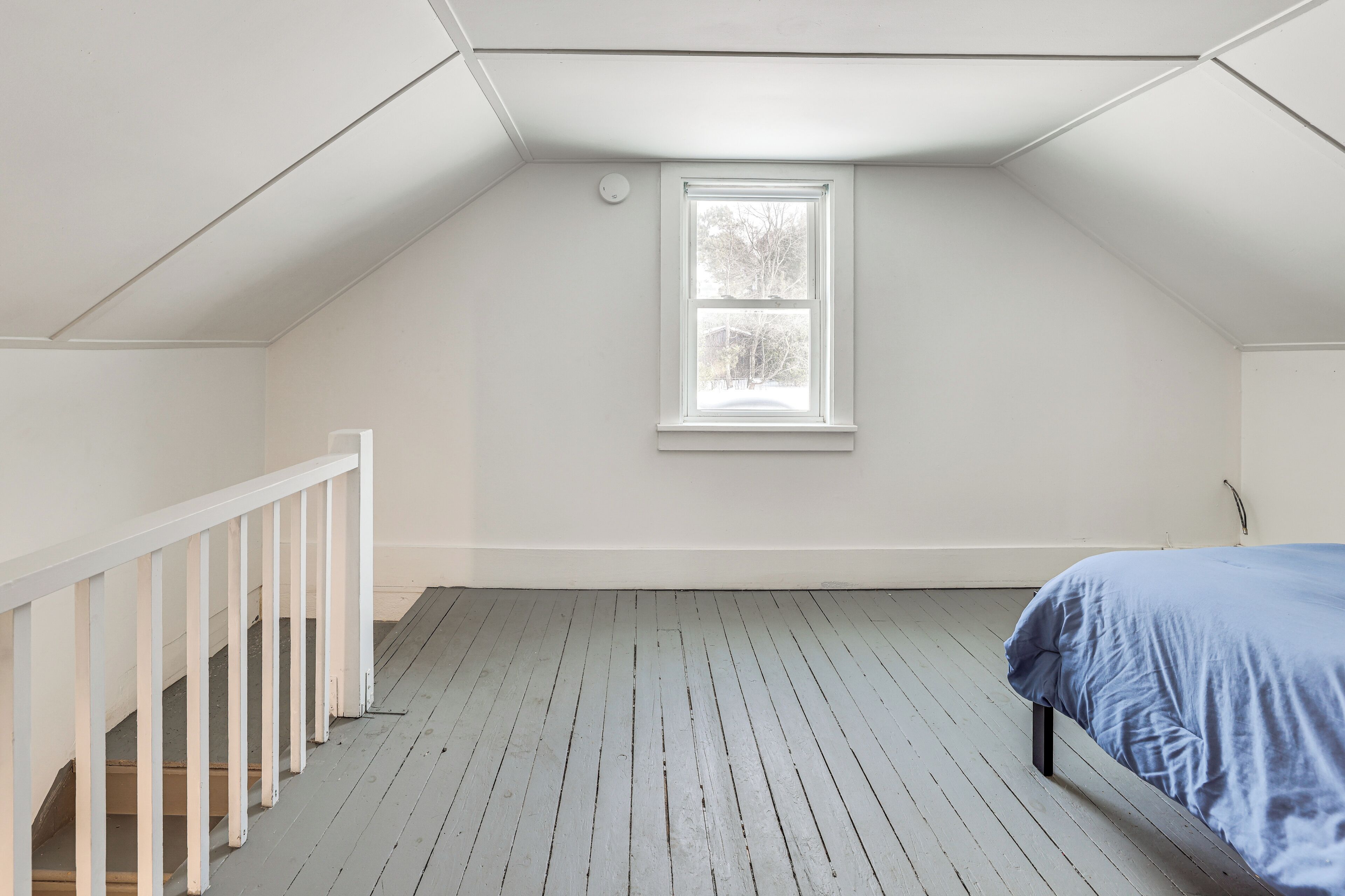 Une pièce avec un plafond en pente, une fenêtre, une rambarde blanche et un lit recouvert d’une couverture bleue.