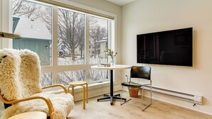 Ein Zimmer mit großem Fenster, einem flachbildfernseher an der Wand, einem gemütlichen Sessel, einem kleinen runden Tisch und einem Stuhl mit Korb.