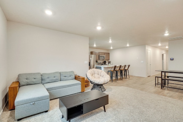 A modern living room with a grey sectional sofa, a black coffee table, and a round chair. A kitchen area with a dining table and chairs is visible in the background.