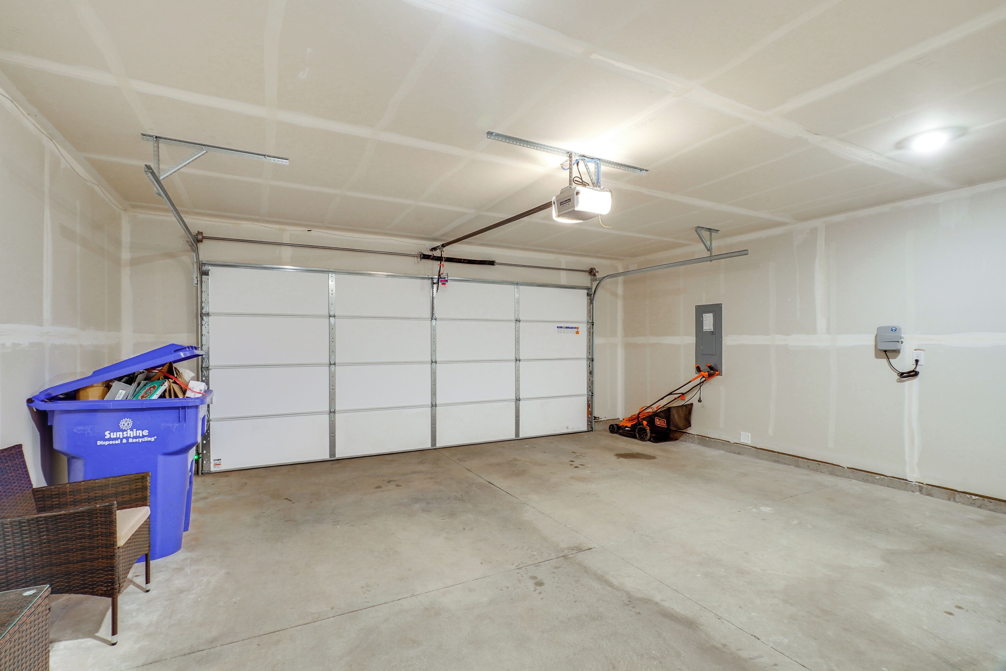 Eine Garage mit geschlossenem Garagentor, einem blauen Müllbehälter mit der Aufschrift 'Sunshine Disposal & Recycling' und einem an der Wand angelehnten Besen.