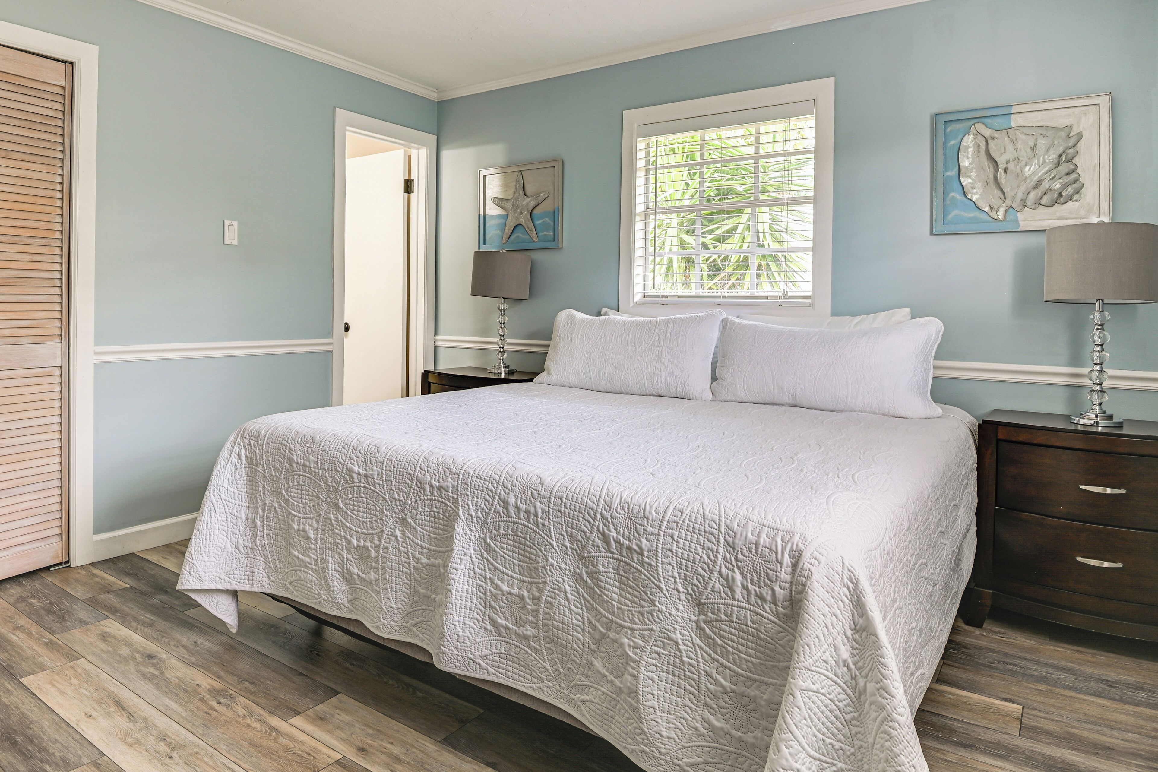 A bedroom with a large bed, a nightstand, a lamp, and a window with blinds.