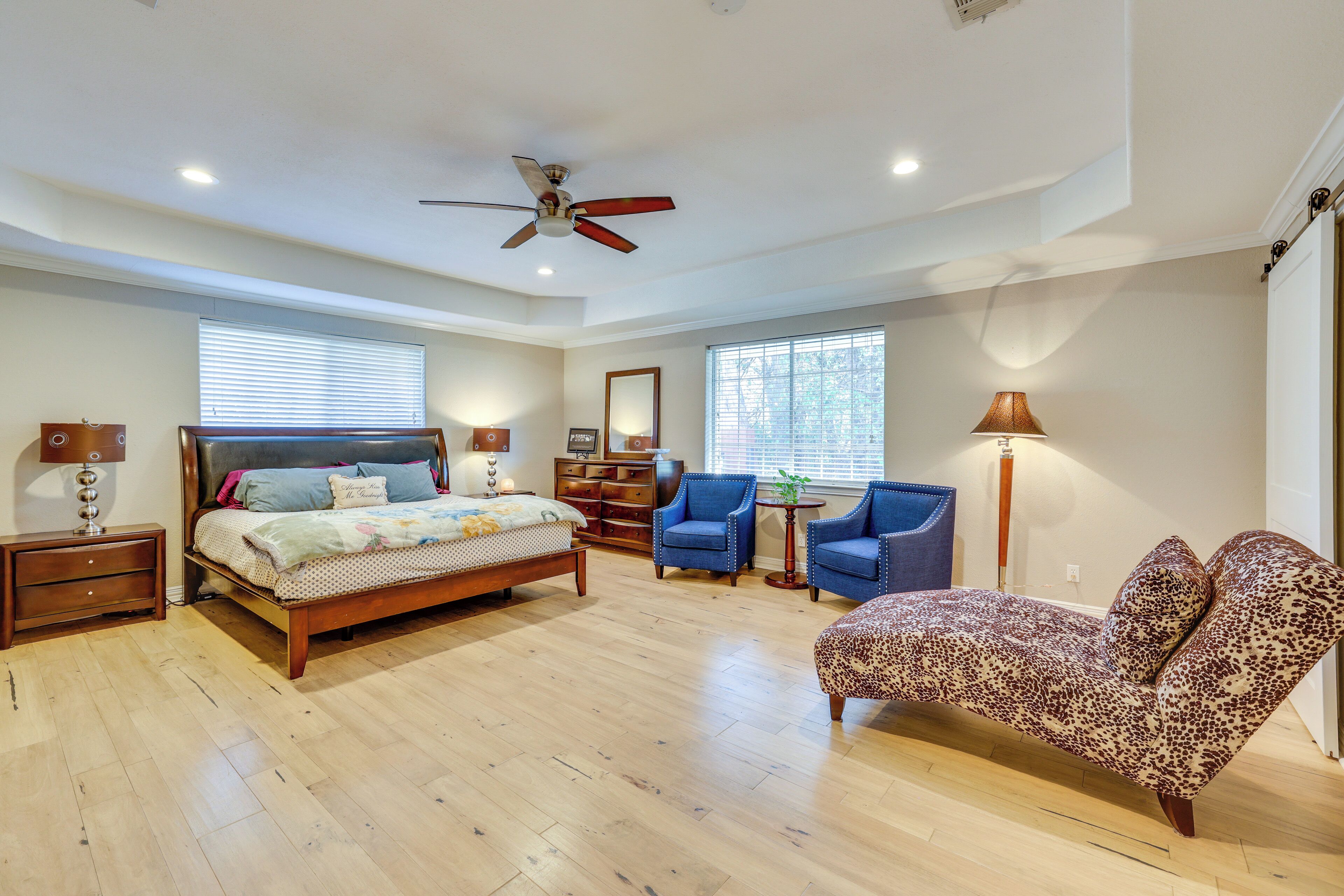 A spacious bedroom with a large bed, two armchairs, a leopard print sofa, a ceiling fan, and a window with blinds.