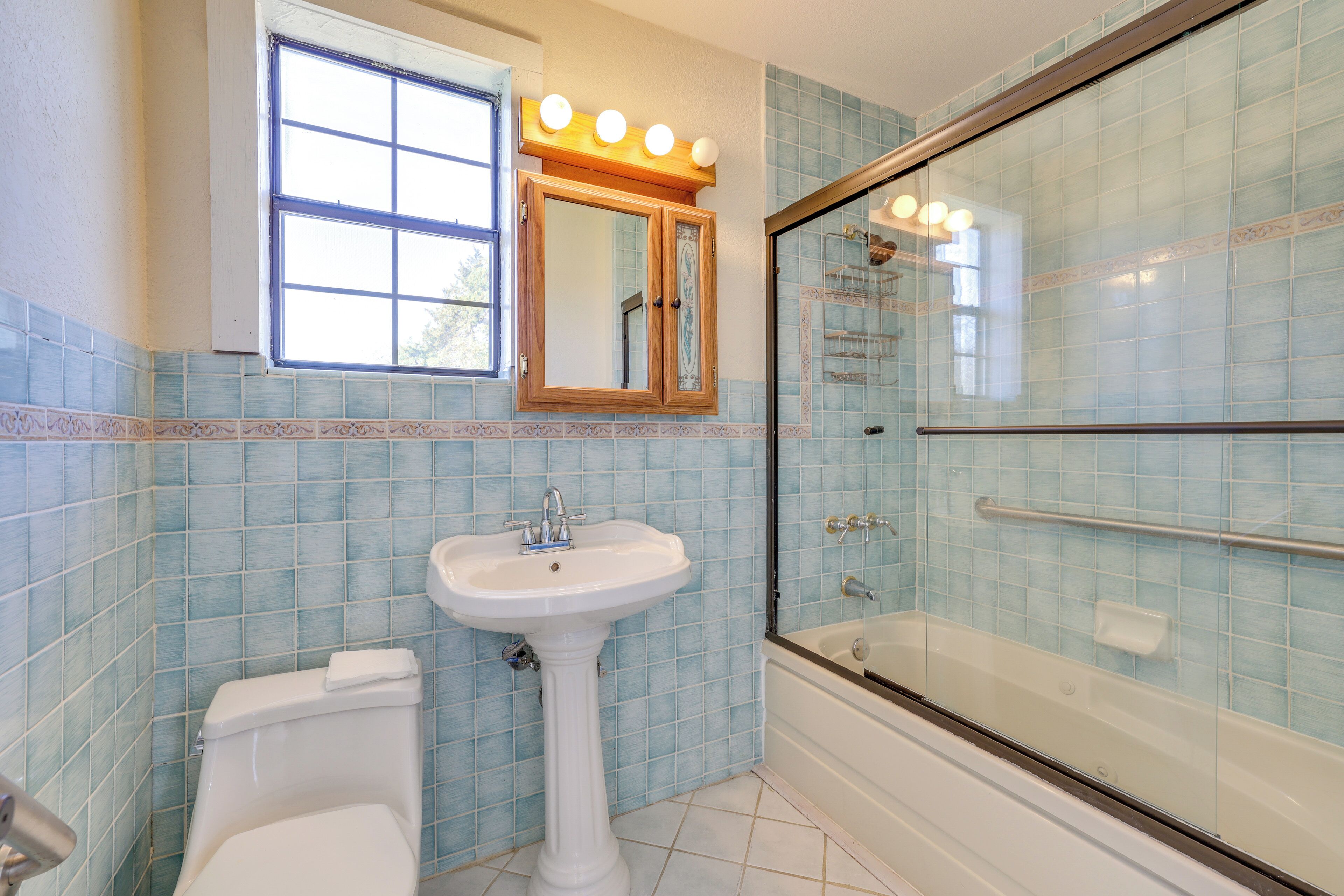 Une salle de bain avec un lavabo sur colonne, des toilettes, une douche à parois de verre et une fenêtre avec des lumières au-dessus du miroir.
