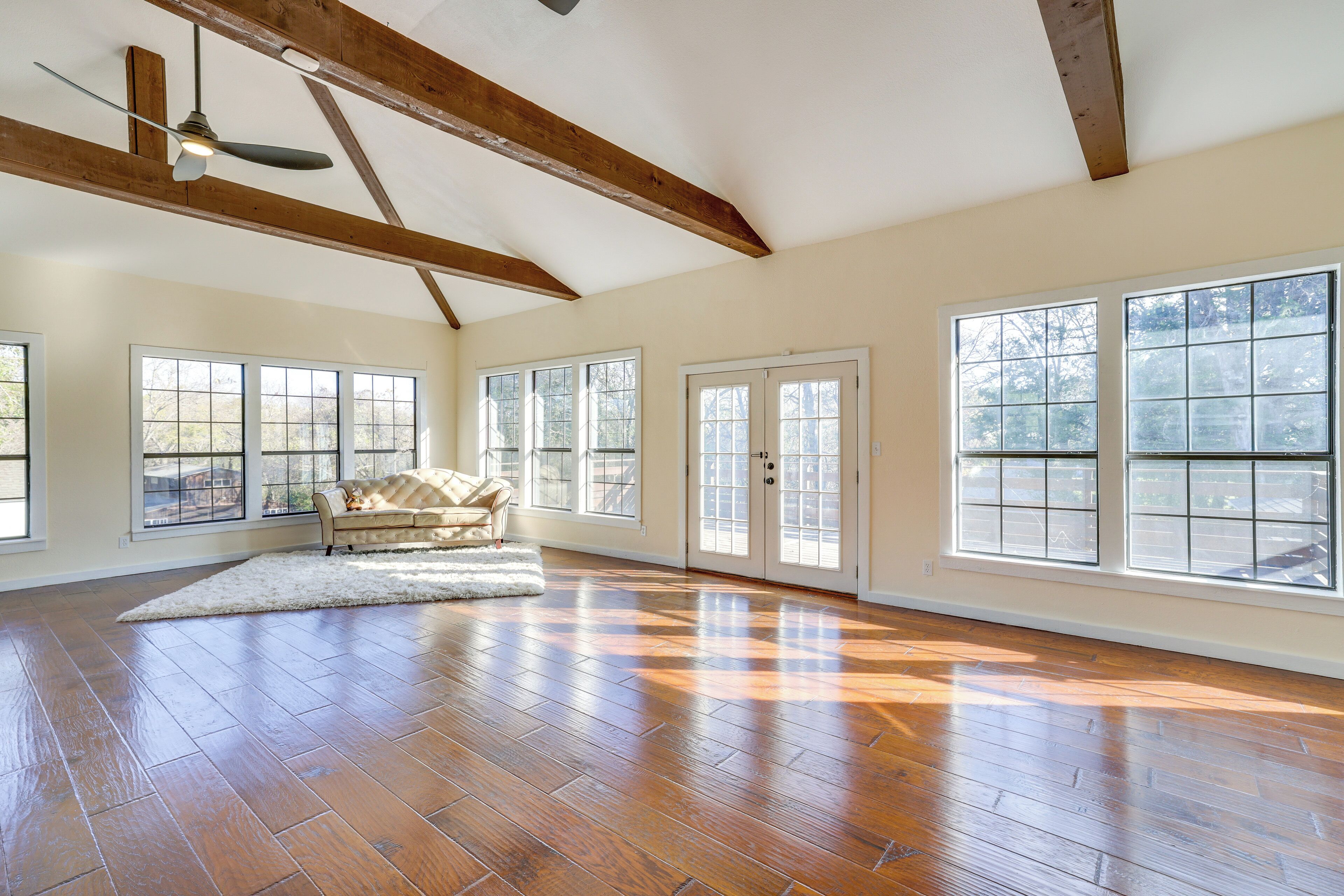 A spacious room with hardwood flooring, large windows, a ceiling fan, and a white rug.