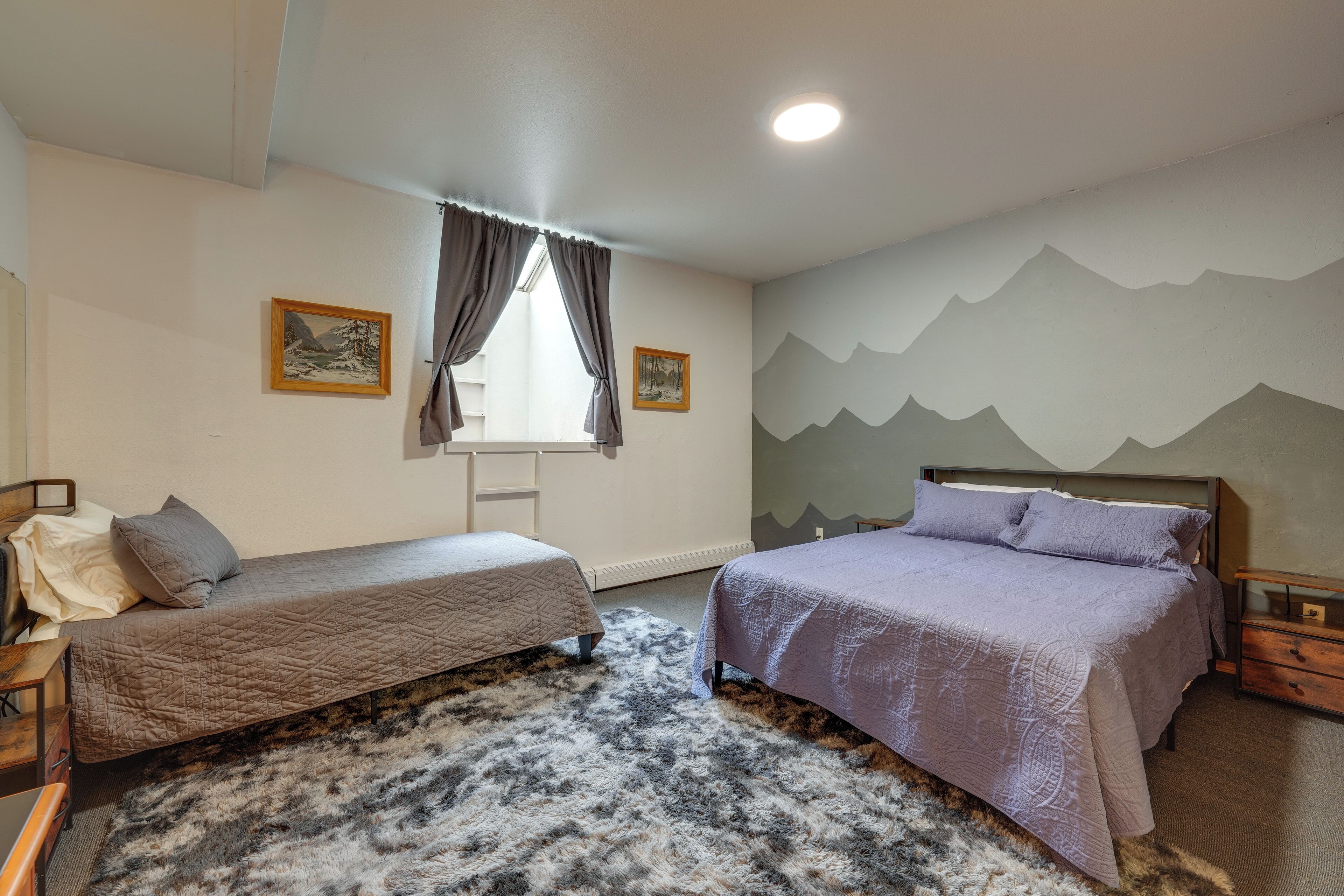 A bedroom with two beds, a window with curtains, a painting on the wall, and a mountain mural.