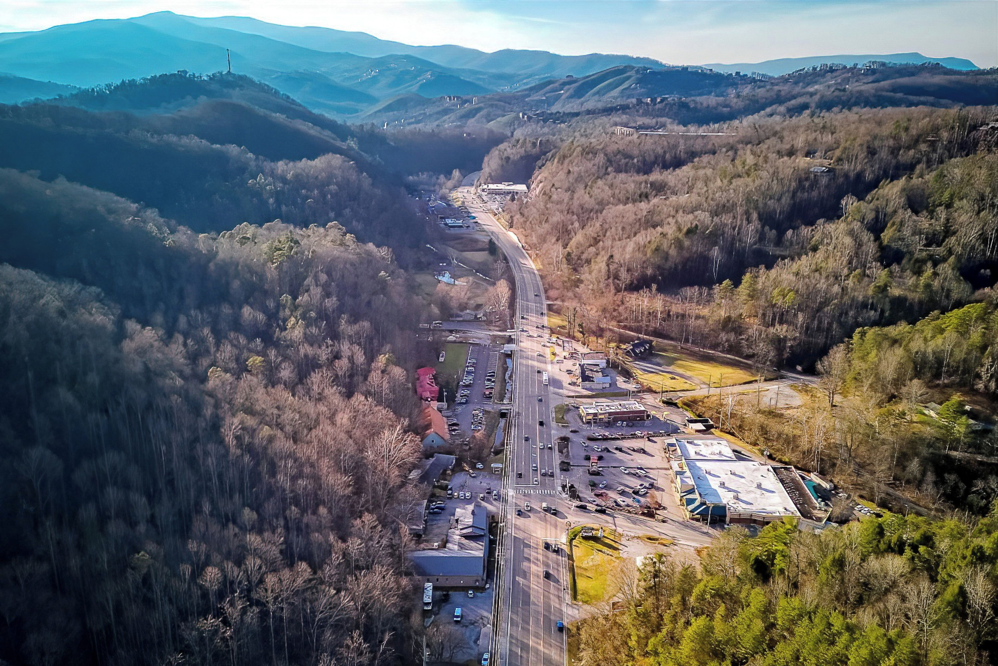 Une vue aérienne d’une autoroute bordée d’un côté et de l’autre par un parking et des bâtiments, le tout entouré de collines boisées.