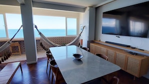 A room with a large table, chairs, a hammock, a TV, and a view of the ocean. - COBAR0108 - Edifício Oceania (Salvador)