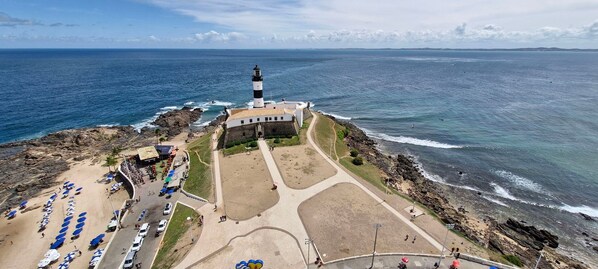 A coastal area with a lighthouse, beach with umbrellas, and a parking lot. - COBAR0108 - Edifício Oceania (Salvador)