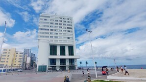 A modern building with a white facade and green shutters, located near a beach with people walking and a boat in the background. - COBAR0108 - Edifício Oceania (Salvador)