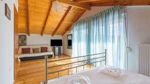 A room with a wooden ceiling, a bed with white linens, a sofa with cushions, a television, and a window with curtains.