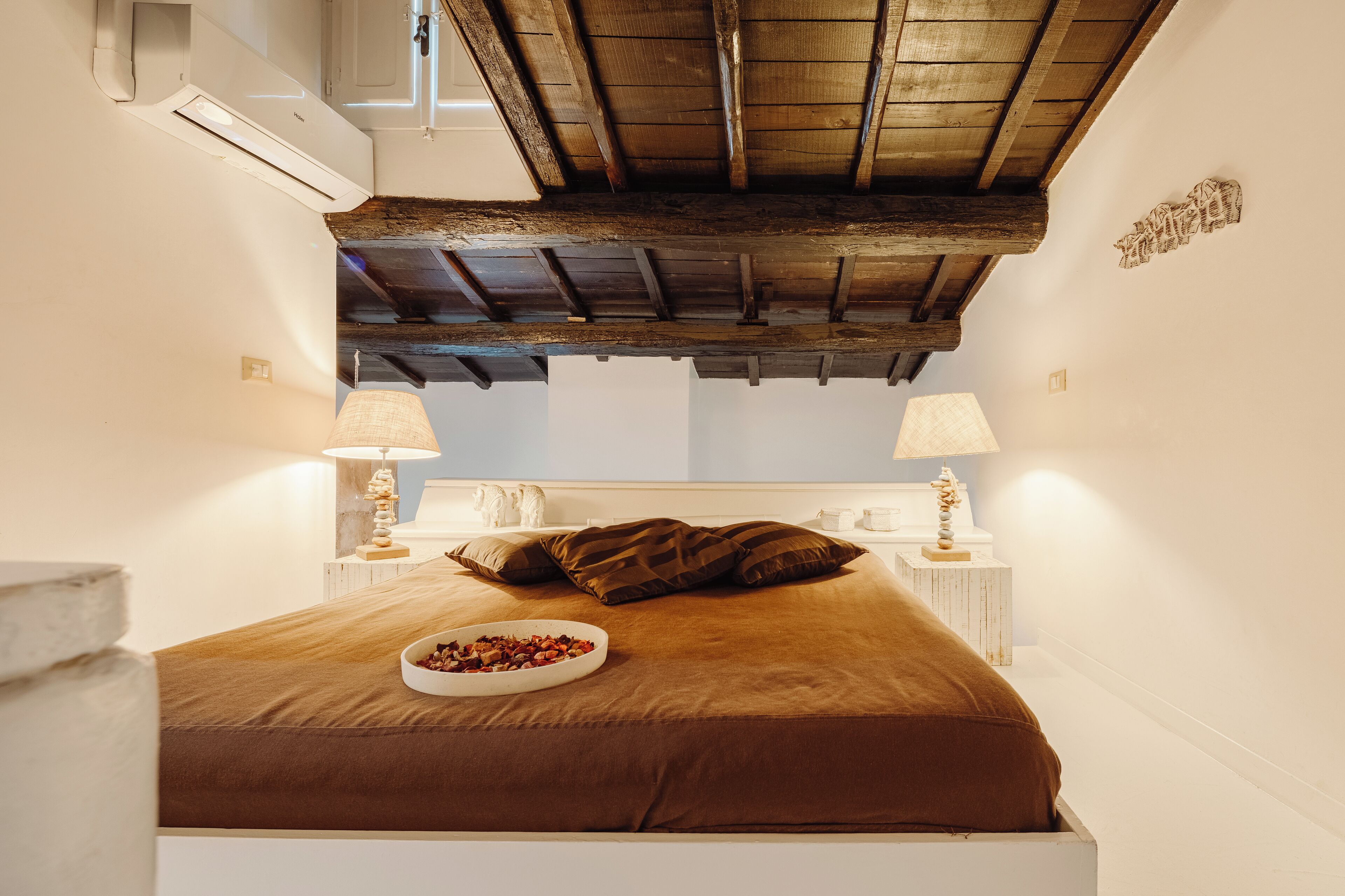 Un dormitorio con techo de madera, una cama con ropa de cama marrón, dos lámparas de noche y un pequeño objeto decorativo sobre la mesita.