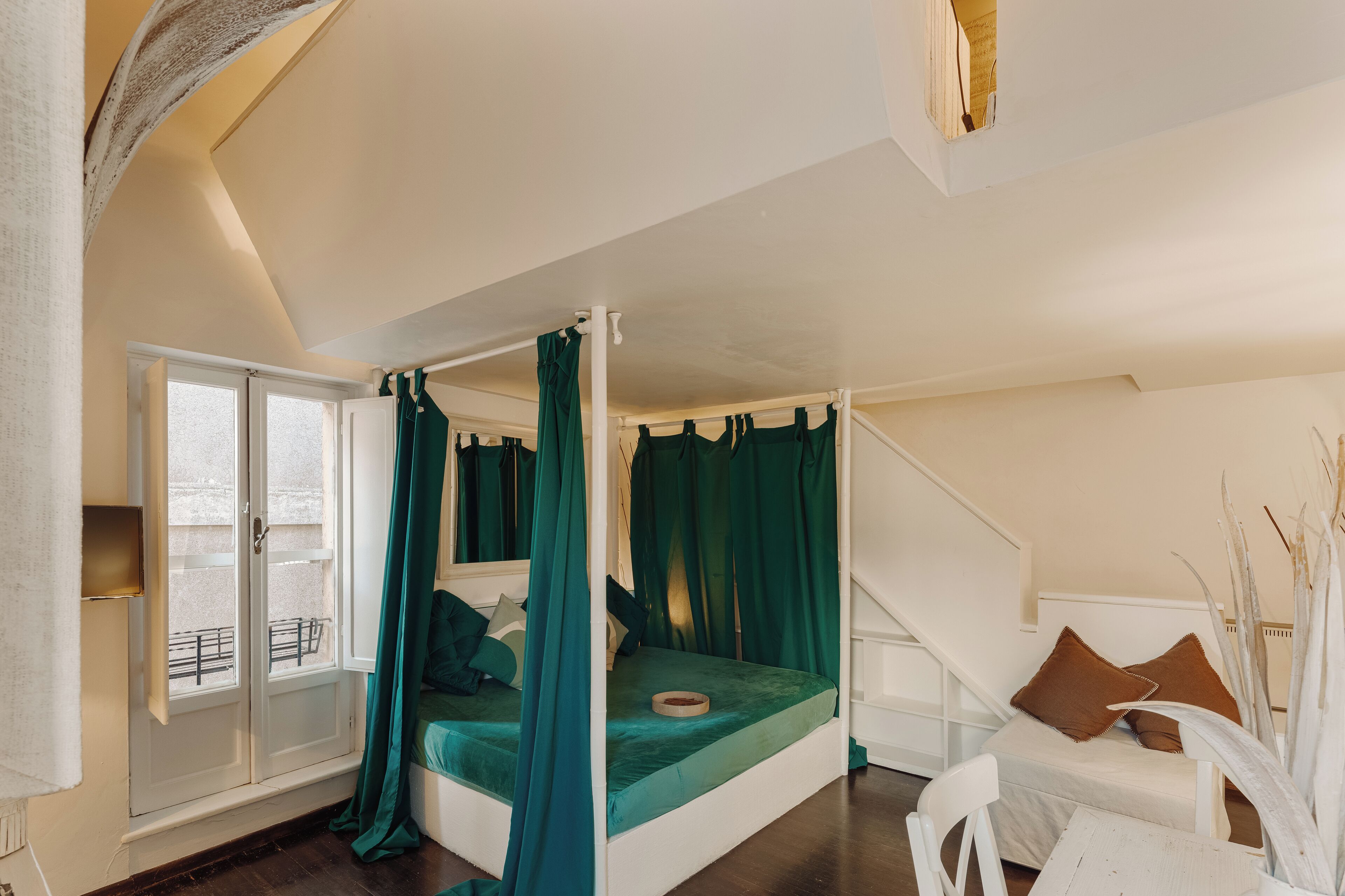 Een bed met hemelbed met groene gordijnen, een witte stoel en uitzicht op een balkon door een schuifdeur.