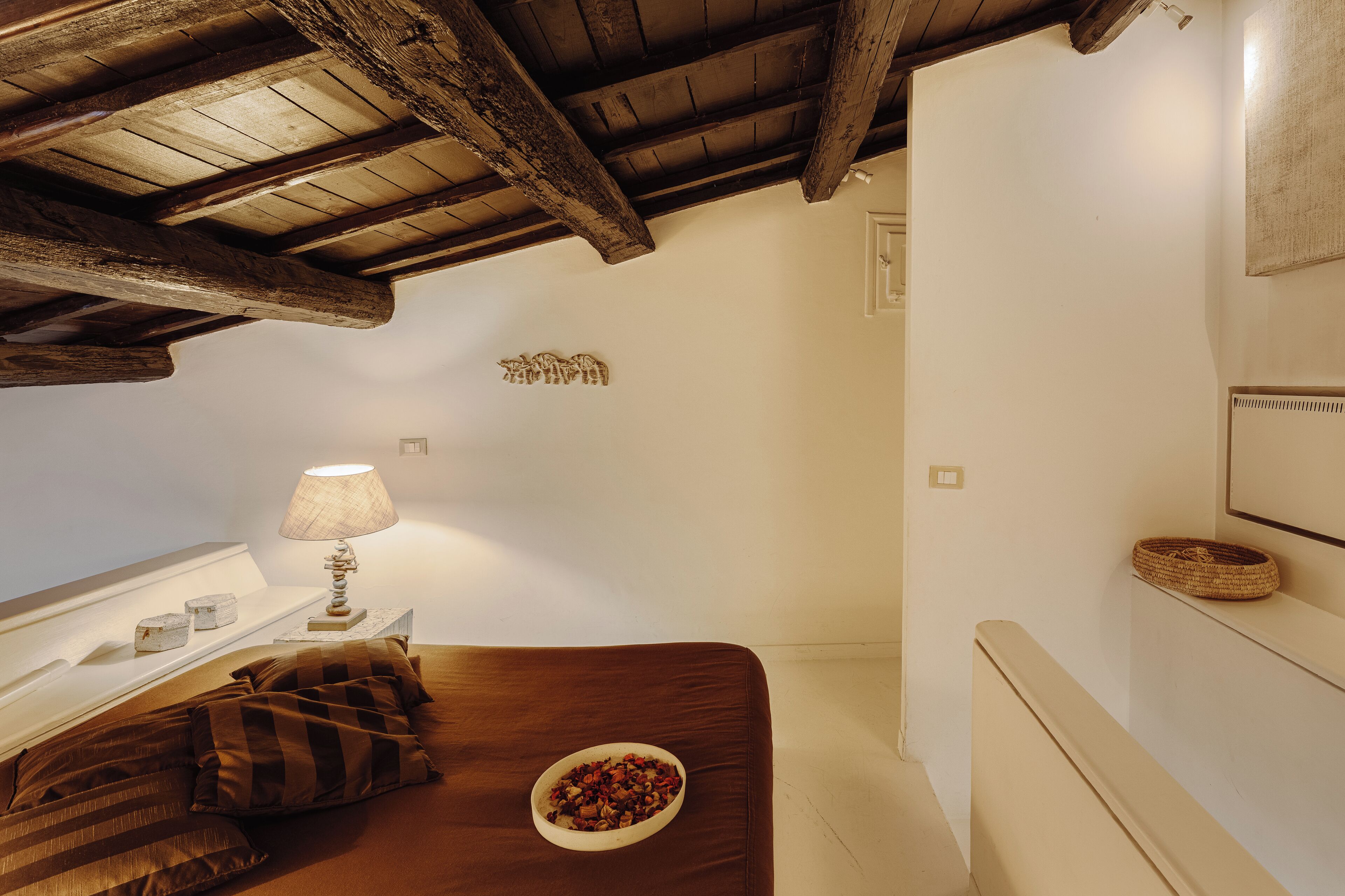 A bedroom with a wooden ceiling, a bed with brown bedding, a bedside table with a lamp, and a small decorative item on the wall.