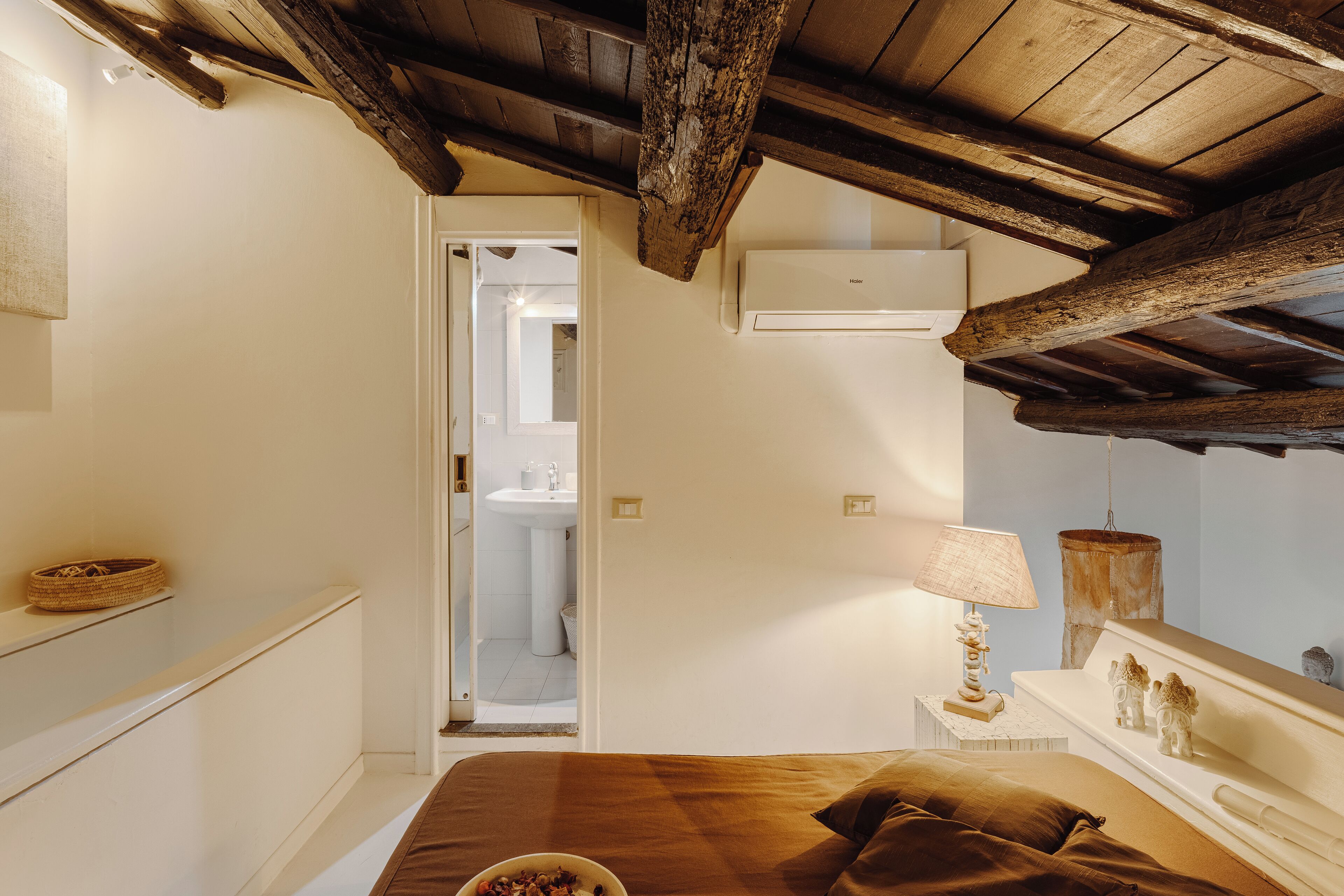 A bedroom with a wooden ceiling, a bed with brown bedding, a bedside table with a lamp, and a small shelf with a basket.