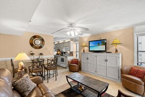 Ein Wohnzimmer mit Sofa, Couchtisch und Fernseher. Es gibt eine Essecke mit Stühlen und Tisch. Der Raum verfügt über einen Deckenventilator und eine Küche im Hintergrund.