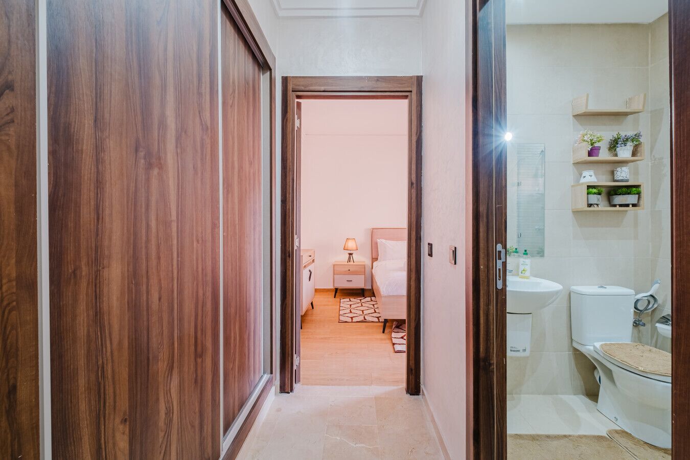 Un corridoio che conduce a una camera da letto con un letto, un comodino e una lampada. C'è un bagno con un lavandino, un water e mensole con piante.