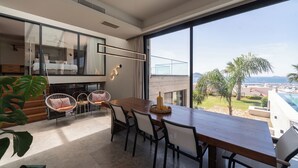 Un espace de repas moderne avec une table et des chaises en bois, de grandes fenêtres offrant une vue sur la mer et les palmiers, et une cloison en verre séparant l’espace de repas de la cuisine.