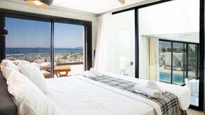 Une chambre d’hôtel avec un grand lit, une vue sur la ville et la mer, une piscine et un balcon équipé d’une table et de chaises.