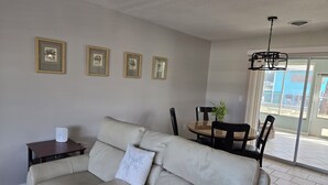 Een woonkamer met een beige bank, een kleine tafel en een eethoek met stoelen en een tafel. Er hangen ingelijste foto's aan de muur en is een plafondlamp.