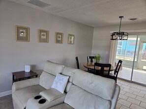 Ein Wohnzimmer mit einem beigen Sofa, einem kleinen Tisch und einem Essbereich mit Stühlen und einem Tisch. An der Wand hängen gerahmte Bilder und eine Deckenleuchte.