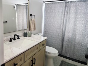 Et badeværelse med en hvid vask, vandhane i sort, beige skab, hvid toilet og bruseforhæng med et sort-hvidt mønster.