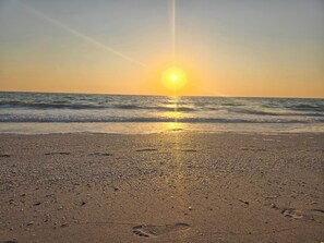 Ein Strand mit Fußabdrücken, die zum Wasser führen, Wellen und einer untergehenden Sonne.
