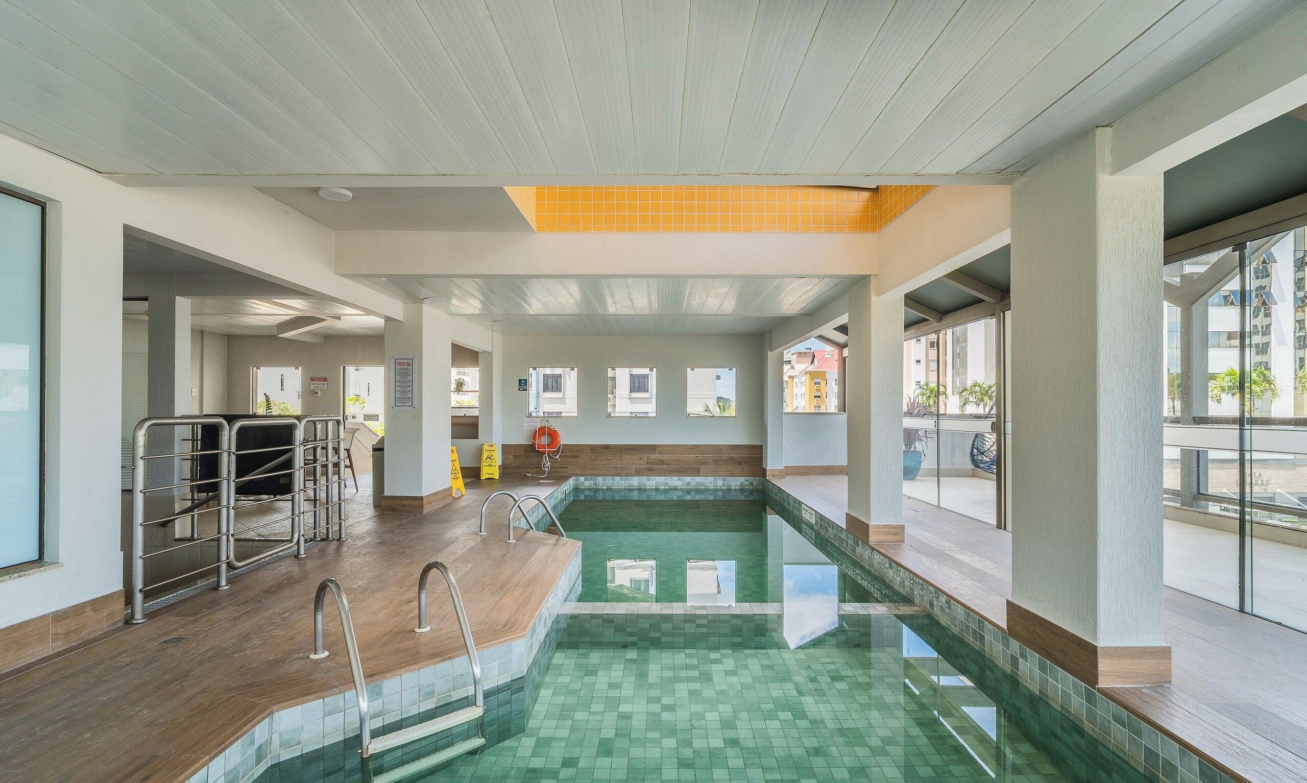 An indoor swimming pool with clear glass walls, metal railings, and a tiled floor.