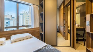 A hotel room with a bed, a large window, a wardrobe, and a view of the cityscape. - Apt em Cond c/ piscina aquecida em Floripa CUR0716 (Florianópolis)