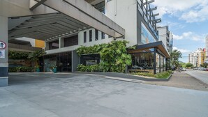 A modern building with a covered entrance, glass windows, and a small outdoor seating area with plants. - Apt em Cond c/ piscina aquecida em Floripa CUR0716 (Florianópolis)