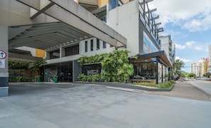 A modern building with a covered entrance, glass windows, and a small outdoor seating area with plants. - Apt em Cond c/ piscina aquecida em Floripa CUR0716 (Florianópolis)