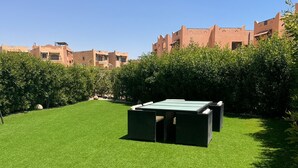 Un espace verdoyant bien entretenu, avec une table et des chaises d’extérieur vertes, entouré d’une végétation dense et avec des bâtiments en arrière-plan.