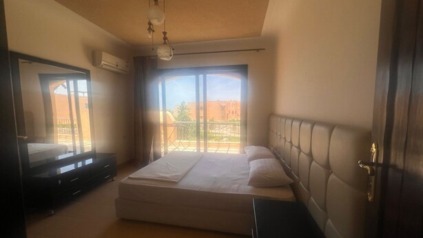 Une chambre à coucher comprenant un lit, une commode, un téléviseur et une fenêtre donnant sur des immeubles et des arbres.