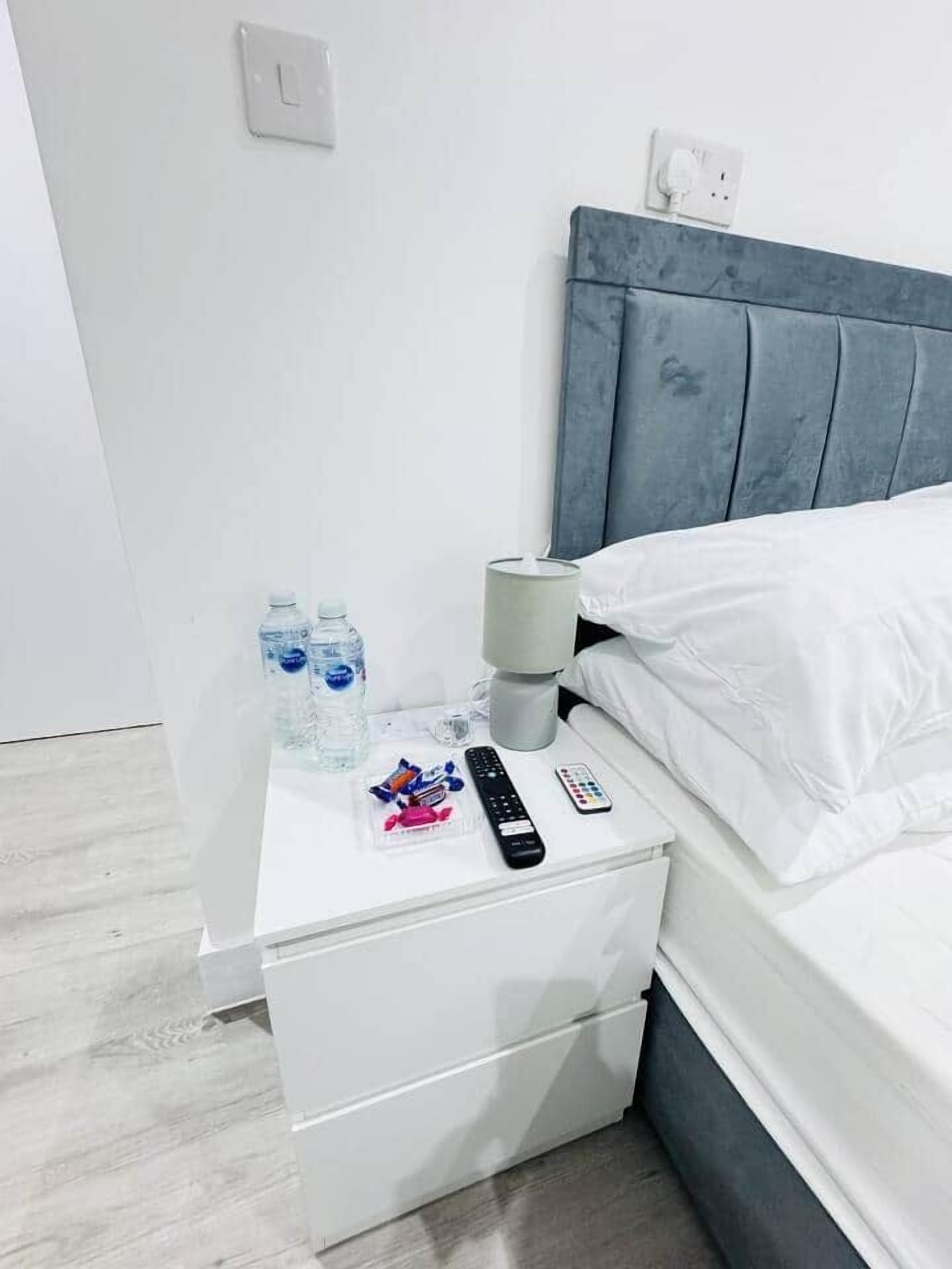 Ett hotellrum med ett vitt sängbord, en grå sänggavel, en vit säng och ett sidobord med en lampa, vattenflaskor, snacks och en fjärrkontroll.