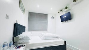 A modern hotel room with a bed, bedside table, water bottles, a lamp, a clock, a TV, and a window with blinds. - Private En-Suite Room near Cardiff City Centre (Cardiff)