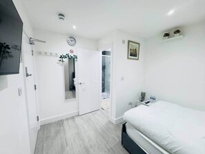 A bedroom with a bed, a nightstand, a clock, a door, and a shower area. - Cosy En-Suite Room near Cardiff City Centre (Cardiff)